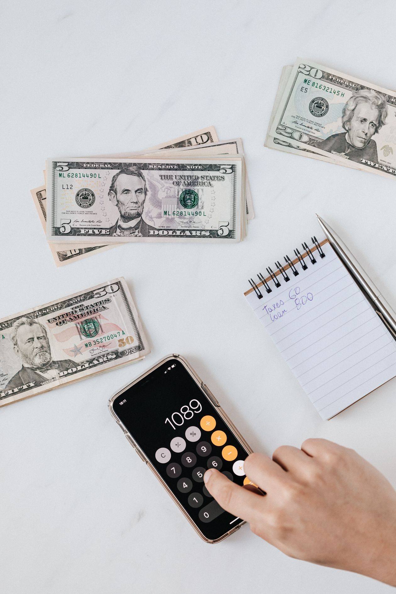 A hand using a calculator beside dollar bills and a notebook while working out a monthly budget